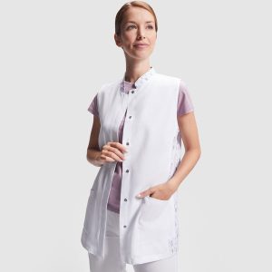 Mujer de pie mirando hacia un lado, vistiendo una bata blanca sin mangas con botones a presión, bolsillos frontales y detalles estampados en los laterales, sobre fondo claro liso.