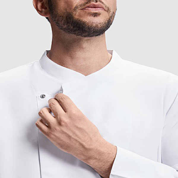 Un hombre lleva la Chaqueta de cocina, de cuello alto y con un bolsillo rectangular en la manga superior, sobre un fondo liso de color gris claro.