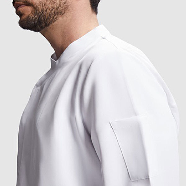 Un hombre lleva la Chaqueta de cocina, de cuello alto y con un bolsillo rectangular en la manga superior, sobre un fondo liso de color gris claro.