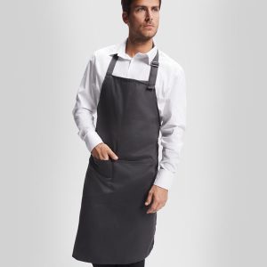 Un hombre con camisa de vestir blanca y un Delantal sobre un fondo liso gris claro, con una mano en el bolsillo del delantal y la otra a su lado: la perfecta ropa de cocina para profesionales.