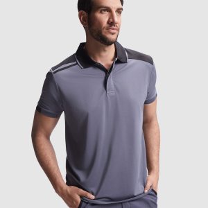 Un hombre de pie con las manos en los bolsillos, mirando hacia un lado, vistiendo un Polo de manga corta antibacteriano gris claro con detalles oscuros en los hombros sobre un fondo claro liso.