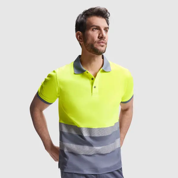 Un hombre de pie con las manos a la espalda, vestido con un Polos de alta visibilidad de color amarillo brillante y gris con rayas horizontales, mira ligeramente hacia un lado sobre un fondo liso de color gris claro.